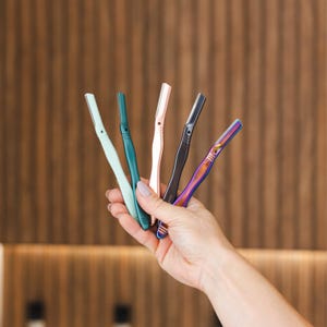 May include: A hand holds five eyebrow razors in various colors: mint green, teal, rose gold, black, and iridescent. Each razor has a blade and a handle. The background is a wooden wall.