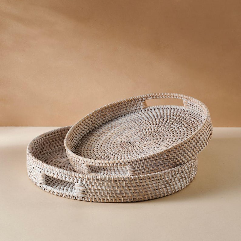 May include: Two white wicker trays with handles. The larger tray is stacked on top of the smaller tray.