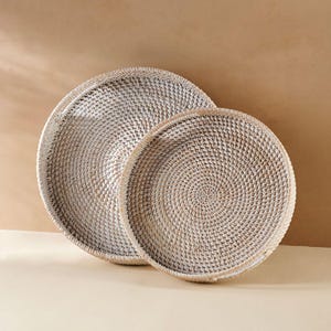 May include: Two white wicker trays, one nested inside the other. The trays are round and have a woven pattern.
