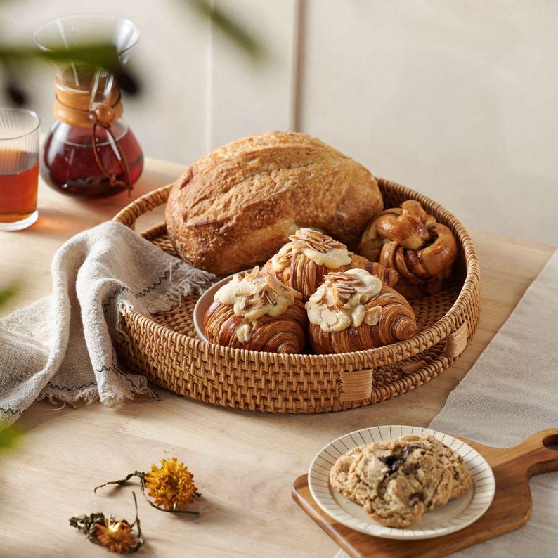 May include: A woven wicker tray with a handle, filled with baked goods. The tray contains a loaf of bread, a cinnamon roll, and three pastries topped with sliced almonds. The tray is sitting on a wooden table.