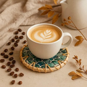 Könnte beinhalten: Eine weiße Keramik-Kaffeetasse gefüllt mit einem Latte, der ein blattförmiges Milchschaummuster aufweist. Die Tasse steht auf einem runden Untersetzer mit grün-blauem Schuppenmuster und hellem Rattangeflecht, umgeben von gerösteten Kaffeebohnen.