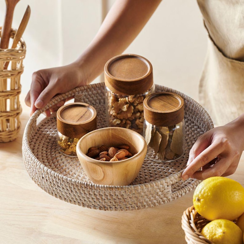 May include: A round woven tray with a natural wicker finish holds three glass jars with wooden lids filled with almonds, walnuts, and bay leaves. A wooden bowl filled with almonds sits in the center of the tray.