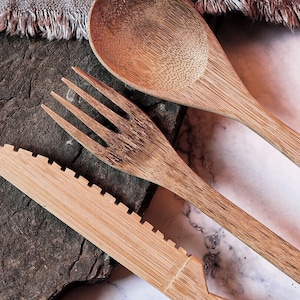Könnte beinhalten: Ein Set aus Holzlöffel, Gabel und Messer. Das Besteck besteht aus hellbraunem Holz und ist auf einer dunkelgrauen Steinoberfläche angeordnet. Das Messer hat eine gezahnte Kante. Im Hintergrund befindet sich ein graues Tuch mit Fransen.