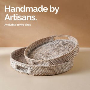May include: Two white woven wicker trays with handles. The trays are round and have a natural woven pattern. The text "Handmade by Artisans. Available in two sizes." is displayed above the trays.