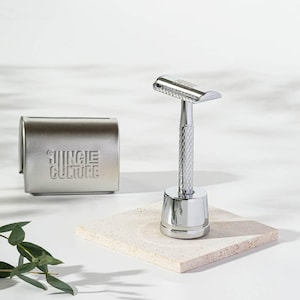 Könnte beinhalten: Ein verchromter Rasierer mit strukturiertem Griff und passendem Ständer. Daneben eine Rasierklingenbox aus Metall mit dem geprägten Text "JUNGLE CULTURE". Die Artikel stehen auf einem hellen Stein.