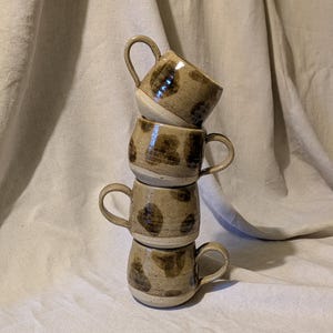 May include: Four handmade ceramic mugs stacked. The mugs are beige with brown spotted patterns and curved handles. The mugs are arranged in a tower formation. The background is a neutral fabric, providing a simple backdrop for the mugs.
