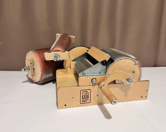 May include: A wooden wool carder with two sets of rotating brushes. The carder has a wooden handle and a metal frame. The carder is labeled "КАНА КОЛЯ" in Cyrillic script.