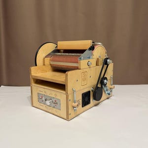 Può includere: Una cardatrice di fibre in legno con un tamburo rosso e beige. La macchina ha un pannello di controllo con una manopola e pulsanti. Una cinghia nera collega il motore al tamburo. La macchina è progettata per la lavorazione delle fibre.