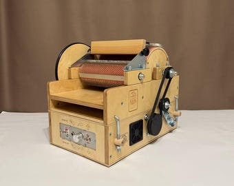 May include: A wooden fiber carding machine with a red and tan drum. The machine has a control panel with a knob and buttons. A black belt connects the motor to the drum. The machine is designed for processing fibers.