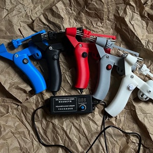 May include: Five tattoo machines in different colours with a black power adapter. The machines are blue, black, red, grey, and white. The power adapter has a digital display and a knob to adjust the power.