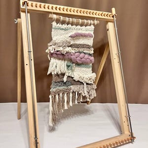 May include: A wooden loom with a tapestry woven on it. The tapestry is made with a variety of colours and textures, including white, brown, pink, and green. The loom is on a stand and has a wooden frame.