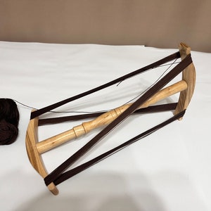 Op de afbeelding: Een houten garenwinder met bruin garen eromheen gewikkeld. De winder heeft een houten spil en wordt ondersteund door vier bruine koorden.