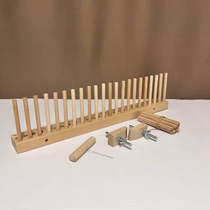 Puede incluir: Un telar de madera con un juego de agujas de tejer de madera, dos abrazaderas de madera, una lanzadera de madera y una aguja de metal. El telar está hecho de madera natural y tiene un color marrón claro.