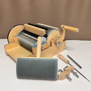 May include: A wooden wool carding machine with a hand crank, metal rollers, and a wooden tray. A hand carder and several tools are also included. The machine is made of light-coloured wood and metal.