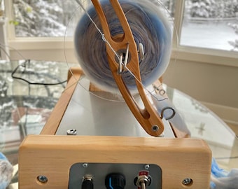 May include: A wooden spinning wheel with a clear plastic shield and a light blue and white yarn. The wheel has a control panel with a black knob and a switch. The background shows a snowy landscape.