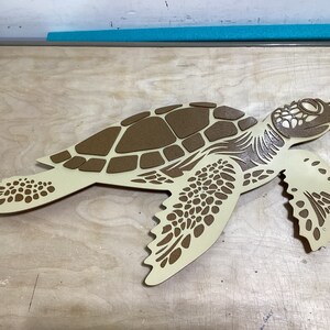 May include: A beige wooden cutout of a sea turtle with a brown shell. The turtle is facing right and has a detailed design.