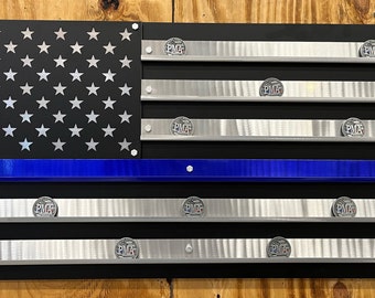 May include: A black and blue metal American flag with a thin blue line, featuring a design for displaying challenge coins. The flag has 13 horizontal stripes, with 50 white stars on a black background. The blue line is centered on the 7th stripe. The flag has 10 rows of silver metal strips for displaying challenge coins. Each row has a small silver coin with the letters "PMF" on it.