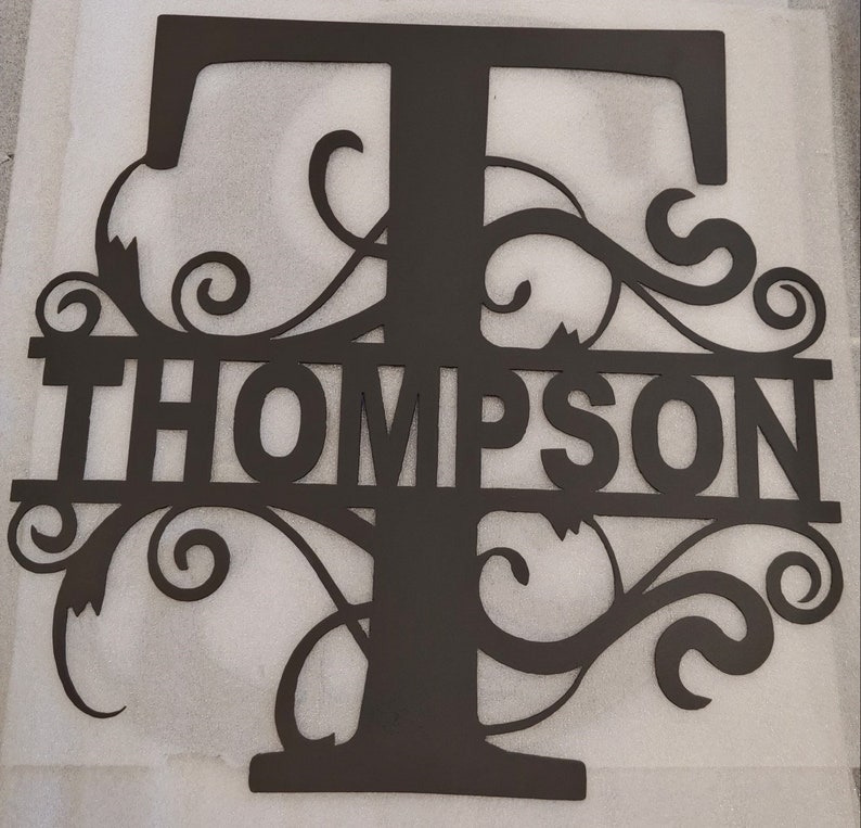 Może przedstawiać: Czarna metalowa dekoracja ścienna z dużą literą T z ozdobnymi zwojami i napisem "THOMPSON" poniżej.