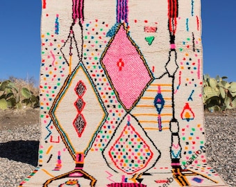 Pode incluir: Um tapete marroquino branco com um padrão geométrico colorido. O tapete apresenta formas de diamantes rosa, azul, verde, laranja e preto sobre um fundo branco. O tapete tem uma borda de franjas.