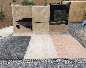 May include: A handwoven rug with a geometric design in black, white, and beige. The rug has a fringe border and is displayed against a backdrop of a stone wall and a hillside.
