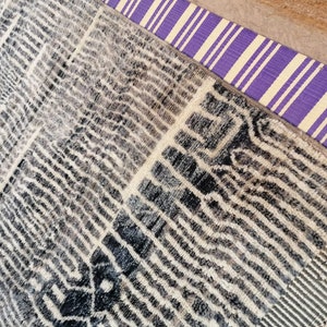 May include: A black and white patterned rug with a purple and white striped border. The rug has a fringe on the bottom edge.