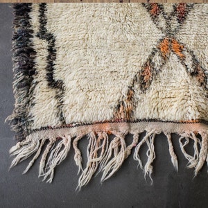 May include: A white, handwoven rug with brown and orange geometric patterns. The rug has a fringe border and is made of wool.