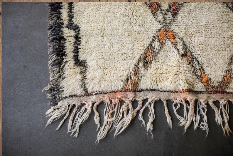 May include: A white, handwoven rug with brown and orange geometric patterns. The rug has a fringe border and is made of wool.