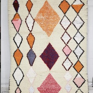 May include: A white rug with a geometric pattern of diamonds in shades of orange, pink, purple, and black. The diamonds are arranged in rows and columns, creating a symmetrical design.