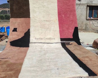 May include: A hand-woven wool rug with a geometric design in shades of brown, pink, white, and black. The rug has a fringe border and is hanging on a wall. The text "Berber Creations" is visible in the center of the rug.