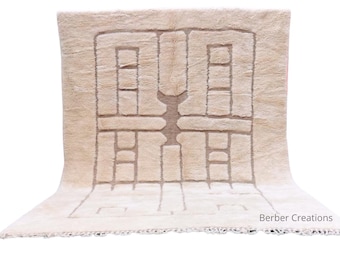 May include: Beige rectangular rug with a geometric design in a lighter beige color. The rug has a central pattern with rectangular and square shapes. The rug has a fringe along the bottom edge. The text "Berber Creations" is in the bottom right corner.