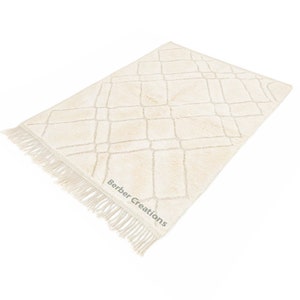 May include: A cream-colored rug with a diamond pattern and fringe. The rug has a subtle texture and is made of a soft material. The rug is labeled "Berber Creations".