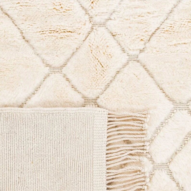 May include: A cream-colored rug with a diamond pattern and fringe. The rug is made of a soft, fluffy material.