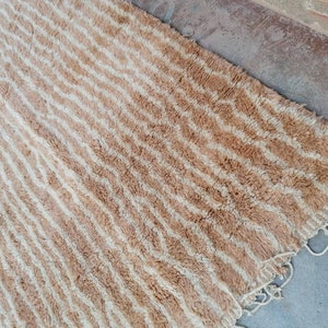 Puede incluir: Una alfombra rayada beige y blanca con un borde de flecos. La alfombra tiene un patr&oacute;n ondulado y est&aacute; hecha de un material suave y esponjoso.