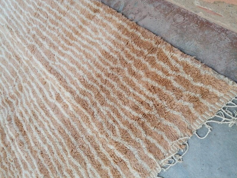 Puede incluir: Una alfombra rayada beige y blanca con un borde de flecos. La alfombra tiene un patr&oacute;n ondulado y est&aacute; hecha de un material suave y esponjoso.