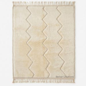 May include: Cream-colored rectangular rug with a zigzag pattern in a lighter shade. The rug has a fringed edge and the text "Berber Creations" in the bottom right corner. The rug is a neutral color and would fit in many home decor styles.