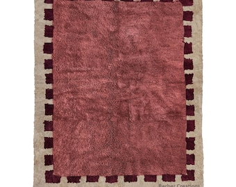 Puede incluir: Una alfombra rectangular con un centro afelpado de color rosa rojizo y un borde beige. El borde presenta un patrón repetitivo de cuadrados rojos oscuros. La alfombra tiene flecos a lo largo del borde inferior y el texto "Berber Creations".