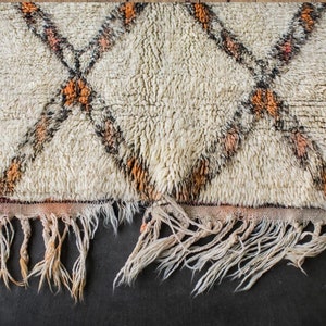 May include: A white rug with a diamond pattern in orange and black. The rug has a fringe border.