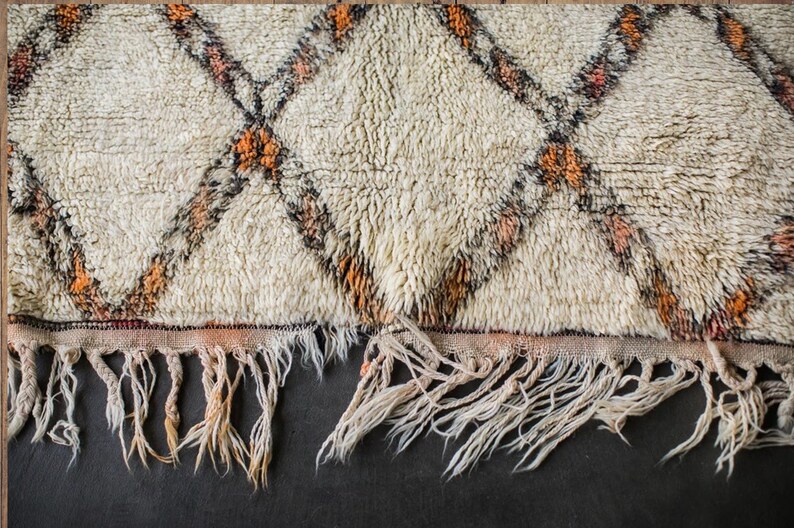May include: A white rug with a diamond pattern in orange and black. The rug has a fringe border.