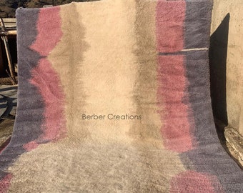 May include: A handwoven Berber rug with a gradient design in shades of pink, beige, and purple. The rug has a fringe border and the text "Berber Creations" is printed in the center.