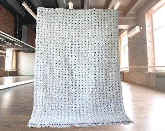 May include: A large, white, textured curtain with a grid-like pattern hangs in a room with a wooden floor and exposed brick walls. The curtain has a decorative, fringed edge with a dark blue trim. The room has large windows and a mirrored wall.