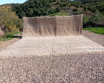 Puede incluir: Una alfombra grande, de color blanco roto, tejida a mano con flecos en todos los lados. La alfombra está extendida sobre una superficie de grava frente a una ladera con árboles.