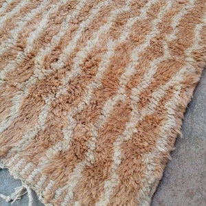 Puede incluir: Una alfombra marroqu&iacute; beige y blanca con un patr&oacute;n geom&eacute;trico. La alfombra tiene un borde de flecos y est&aacute; hecha de lana.