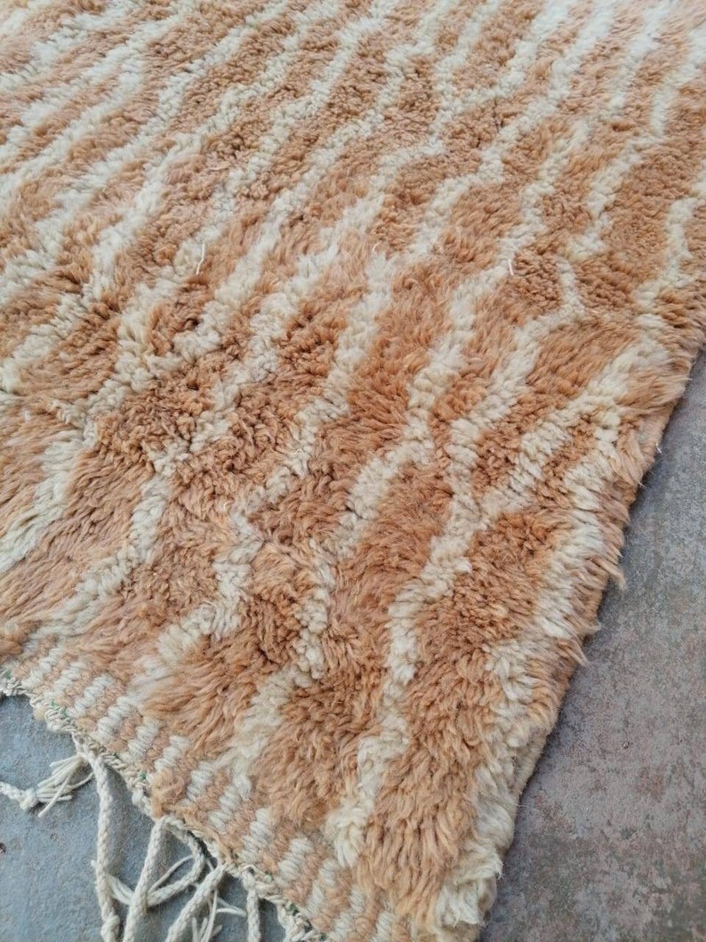 Puede incluir: Una alfombra marroqu&iacute; beige y blanca con un patr&oacute;n geom&eacute;trico. La alfombra tiene un borde de flecos y est&aacute; hecha de lana.