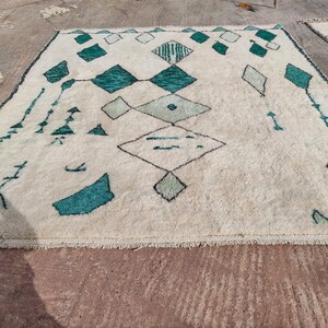 May include: A white rug with a geometric pattern in shades of green. The rug has a fringe border and is made of wool.