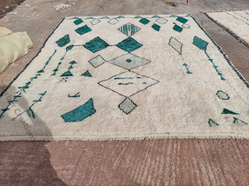 May include: A white rug with a geometric pattern in shades of green. The rug has a fringe border and is made of wool.