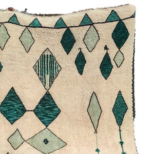 May include: A cream-colored rug with a geometric pattern of green and white diamonds. The rug has a fringe border.