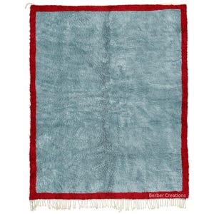 Könnte beinhalten: Ein rechteckiger Teppich mit einem hellblauen Zentrum und einem leuchtend roten Rand. Der Teppich hat eine zottelige Textur und einen Fransenbesatz am unteren Rand. Der Text "Berber Creations" befindet sich unten rechts.