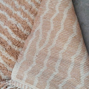 Puede incluir: Primer plano de una alfombra de rayas beige y blancas con textura esponjosa. La alfombra tiene flecos en el borde inferior.