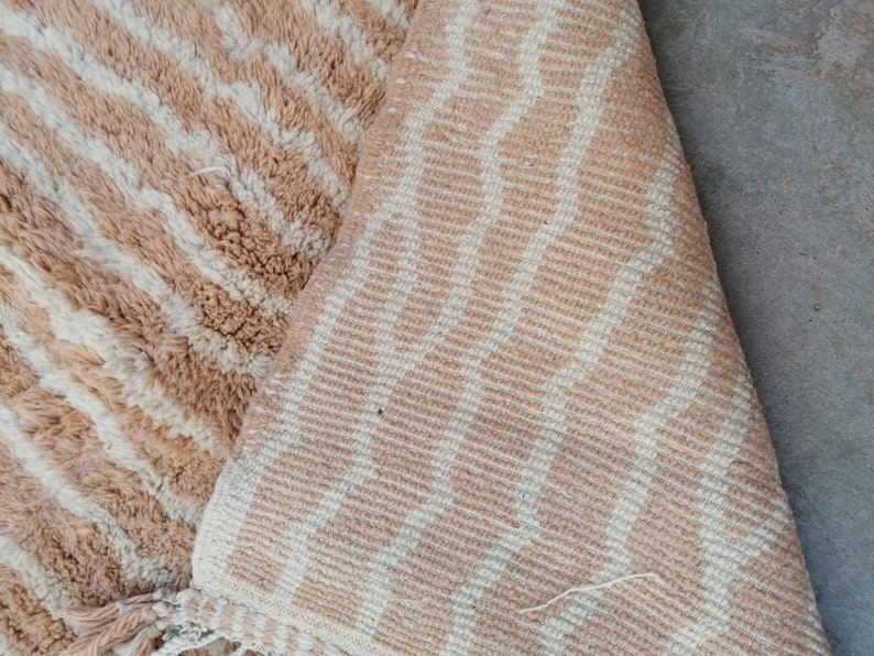 Puede incluir: Primer plano de una alfombra de rayas beige y blancas con textura esponjosa. La alfombra tiene flecos en el borde inferior.