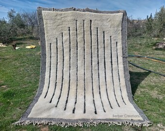Könnte beinhalten: Ein weiß-schwarz gestreifter Teppich mit Fransen. Der Teppich hängt an einer Wäscheleine auf einem grasbewachsenen Feld. Der Text "Berber Creations" ist in der rechten unteren Ecke des Teppichs sichtbar.